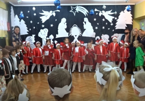 Wszystkie przedszkolaki na tle bożonarodzeniowej dekoracji wszystkie dzieci stoją i trzymając się za ręce śpiewają pastorałkę.
